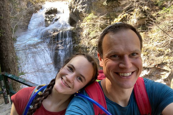 father and daughter smile for the camera in front of a small waterfall in the pyrenees